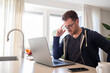 © erikrous - working at home in the kitchen. caucasian male sitting at the kitchen counter behind his laptop working and communicating. Dressed in a hoodie, wearing glasses.
