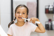 © Sofiia Shunkina - Little Caucasian girl with missing front teeth brushing teeth with electric toothbrush, smiling in bathroom mirror, building healthy habits.