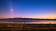 © Elena - Beneath a starry sky, the Milky Way reflects in a calm lake while a wooden fence adds depth to the peaceful landscape at twilight near distant mountains