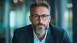 © JoxyAimages - A confident, mature businessman wearing a dark blue suit and glasses is looking directly at the camera in a modern office, reflecting determination and focus.