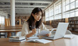 © RooftopStudioBangkok - focused Asian university student studies in library, surrounded by books and laptop. She takes notes and reviews materials, embodying dedication and concentration