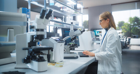 Leinwandbilder - Medical Research and Development Laboratory: Beautiful Caucasian Female Scientist Working on a Computer and Looking at a Petri Dish Sample Under a Microscope. Advanced Biotechnology Lab