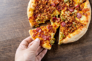 Wall Mural - A hand reaches for a slice of delicious pepperoni and green pepper pizza, featuring crispy toppings, on a rustic wooden background. Perfect for food-related themes or culinary promotions.