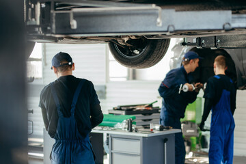 Fototapete - Young car mechanics at repair service station inspecting car wheel and suspension detail of lifted automobile