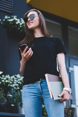 Wall Mural - Positive female student in sunglasses holding smartphone device trying connect to wireless 4g internet while looking away, successful hipster girl with textbook standing outdoors and using cellphone