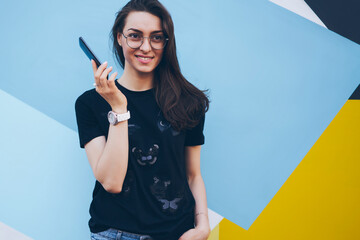 Wall Mural - Smiling fashionable hipster girl in eyeglasses for vision correction standing near colorful background with modern smartphone gadget in hand, positive woman holding mobile phone near bright wall