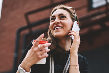 Wall Mural - Young successful female student calling to friend via mobile phone using good 4g internet in roaming, positive smiling woman laughing from joke during international cellular conversation outdoors