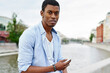 © BullRun - Portrait of african american young man dressed in stylish casual wear looking at camera while sending sms message on smartphone spending leisure time in modern city with river on background