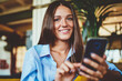© BullRun - Portrait of happy cheerful hipster girl smiling and looking at camera while holding smartphone gadget connected to wireless internet, young successful woman enjoying communication on mobile phone
