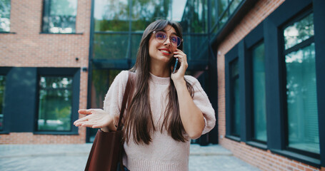 Front camera view of cute Caucasian woman walking outside. Talking on smartphone with friend or relative. Making hand gestures. Wearing round glasses and handbag. Going for stroll in city.