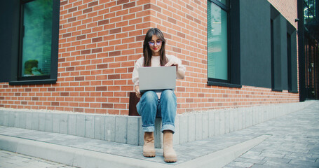 Beautiful Caucasian woman sitting at corner of building. Wearing warm clothing and glasses. Using laptop. Typing on keyboard. Smiling while communicating with someone. Searching web for information.