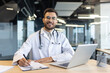© Liubomir - A professional doctor with a stethoscope sits at a desk, smiling confidently while working on paperwork and using a laptop in an office setting.