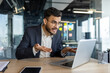 © Liubomir - An exasperated businessman in an office uses a laptop for a video call, expressing frustration. His smartphone is visible, adding to the tech-focused environment.