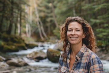 Wall Mural - Portrait of a happy caucasian woman in her 30s dressed in a relaxed flannel shirt while standing against tranquil forest stream