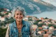 © Markus Schröder - Portrait of a smiling caucasian woman in her 60s wearing a rugged jean vest over picturesque seaside village
