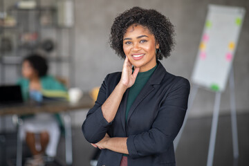 Leinwandbilder - A confident woman stands in a contemporary office, smiling while showcasing teamwork. Other team members engage in discussions at a nearby table, reflecting a cooperative atmosphere.