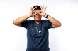 © ArvStd - Impressed funny and cute young Asian man nurse wearing blue uniform with stethoscope looking through fingers over her eyes as if staring in binoculars amazed isolated on white background
