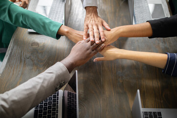 Leinwandbilder - Diverse team members gather around a table in a corporate office, joining hands in a show of cooperation and commitment to success during a productive business meeting.