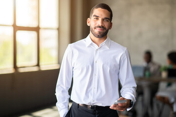 Leinwandbilder - A confident man in a white shirt holds a smartphone while interacting with his diverse team in a corporate office. The atmosphere is focused on teamwork and achieving success during their meeting.