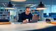 © stock1 - A middle-aged man sits at an office desk, wearing glasses and holding a cup of coffee while working on a laptop. The modern interior design features white furniture and colorful bookshelves