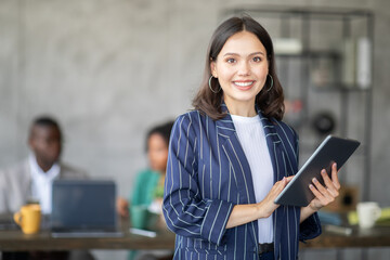 Leinwandbilder - A confident woman stands in front of her diverse team in a contemporary office. She holds a tablet and smiles, as colleagues engage in a productive discussion while working together on projects.