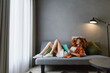© SHOTPRIME STUDIO - Relaxed young woman in casual outfit lounging on a modern couch in a cozy living room, enjoying a book, with a gray backdrop and natural light