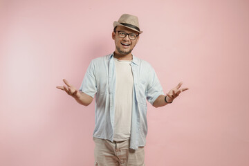 Wall Mural - Cheerful young Indonesian man wearing blue shirt and hat doing welcoming gesture.