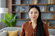 © Liubomir - Asian woman with long hair sitting on sofa at home, exuding confidence and calmness. Background bookshelf filled with books contributes to cozy ambiance and inviting atmosphere.