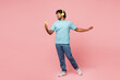 © ViDi Studio - Full body young Indian man wear blue t-shirt casual clothes listen to music in headphones hold use mobile cell phone isolated on plain pastel light pink background studio portrait. Lifestyle concept.