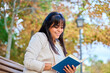 © Oscar - Smiling Latina woman reading a book while sitting on a bench in an autumn park