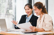 © kenchiro168 - Two professional women discussing work while reviewing documents and using laptop in bright office space, showcasing collaboration and teamwork