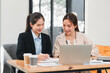 © kenchiro168 - Two professional women collaborating at desk with laptop, documents, and coffee cups, showcasing teamwork and productivity in modern office
