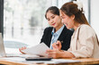 © kenchiro168 - Two professional women discussing documents at desk in modern office, with laptop and papers, showcasing teamwork and collaboration in business