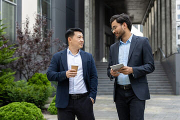 Poster - Two professional men in business attire engage in a friendly discussion while holding coffee cups and tablet. The setting is urban, with greenery and modern architecture in the background.