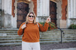 © mdyn - Happy Tourist girl walking in the city during vacation. Cheerful 30s woman in sunglasses pointing finger somesing. Travel concept, old town square. Smiling model posing in the street