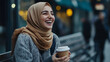 © Anastasia - A middle eastern woman in her 20s smiles joyfully while holding a coffee cup, dressed warmly in a cozy sweater and hijab, sitting outdoors in a busy urban setting