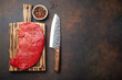© somegirl - Raw uncooked top round beef steak on wooden cutting board with big kitchen knife and pepper on dark brown rustic stone background top view, cooking meat steak with copy space