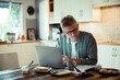 © Davor - Worried senior man reading paperwork while working on laptop at kitchen desk