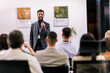 © Mediteraneo - Young business man lecturing his multiethnic colleagues. Having a speech while holding a microphone. Everyone is listening and some are raising their hand.