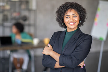 Leinwandbilder - A confident businesswoman stands with arms crossed, smiling in a modern office. In the background, a colleague works at a desk, showcasing a collaborative environment focused on success.