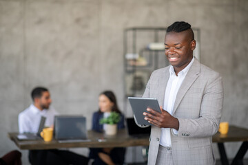 Leinwandbilder - A business professional stands in a stylish office, reviewing information on a tablet while colleagues engage in discussion nearby. The atmosphere highlights teamwork and international cooperation.