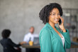 © Prostock-studio - A professional woman in a green blazer smiles as she speaks on the phone at a contemporary office. Team members collaborate in the background, showcasing a diverse work environment.