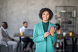 © Prostock-studio - A diverse group of professionals collaborates in a spacious office. One woman in a green blazer holds a tablet, smiling at the camera while her colleagues work together in the background.