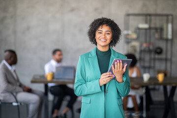 Leinwandbilder - A diverse group of professionals collaborates in a spacious office. One woman in a green blazer holds a tablet, smiling at the camera while her colleagues work together in the background.