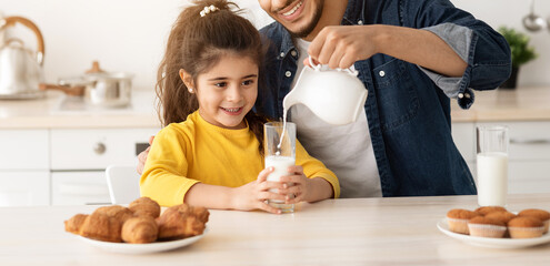 Wall Mural - Snack Time. Happy Arab Dad And Daughter Eating Pastry And Drinking Milk In Kitchen, Cheerful Middle Eastern Daddy And Child Having Fun Together, Caring Father Pouring Calcium Drink From Jug To Glass