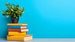 © Chinawat - Books and Plant on Blue Background: A stack of colorful books, each with a distinct spine, sits on a table, a vibrant potted plant adds a touch of life. The composition is simple yet elegant.