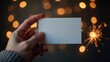 © Hanna - Hand holds blank of white business card on a dark with golden shiny lights background. New year or Christmas gift card certificate. Plain call-card mockup template. A paper namecard. Party invitation