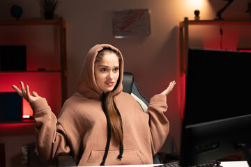 Young beautiful woman programmer in hoodie working overtime at night using pc at home, with surprised expression on her face spread her arms I don't know I'm sorry