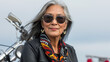 © Creative Content  - A graceful older woman with gray hair in a black leather jacket and scarf, smiling confidently in front of a motorcycle on a cloudy day.
