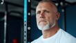 © Iona - A strong, older man with a muscular build stands confidently in a modern gym, embodying strength, dedication, and focus against a backdrop of gym equipment.